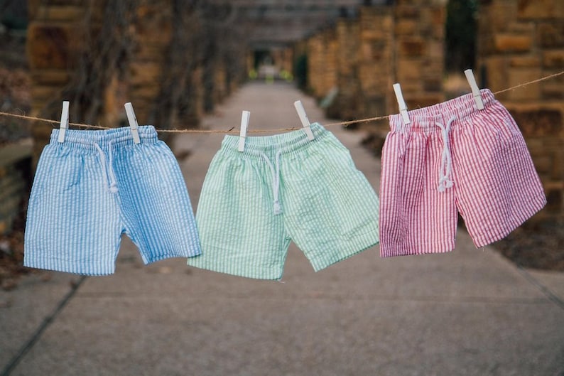 Puede incluir: Tres pares de pantalones cortos de seersucker colgados en un tendedero, cada uno con una cintura de cord&oacute;n blanco. Los pantalones son de color azul claro, verde menta y rosa, con un patr&oacute;n de rayas verticales y acabado de tela texturizada.