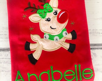 May include: Red Christmas-themed t-shirt with an embroidered image of a reindeer wearing a green bow and a jingle bell necklace. The name "Anabelle" is embroidered in green below the reindeer.