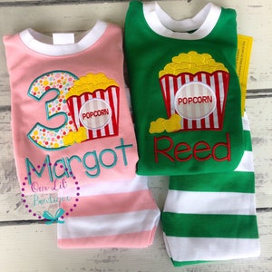 May include: Two pairs of pajamas with a popcorn theme. One pair is pink with white stripes and says "Margot" and "3". The other pair is green with white stripes and says "Reed".