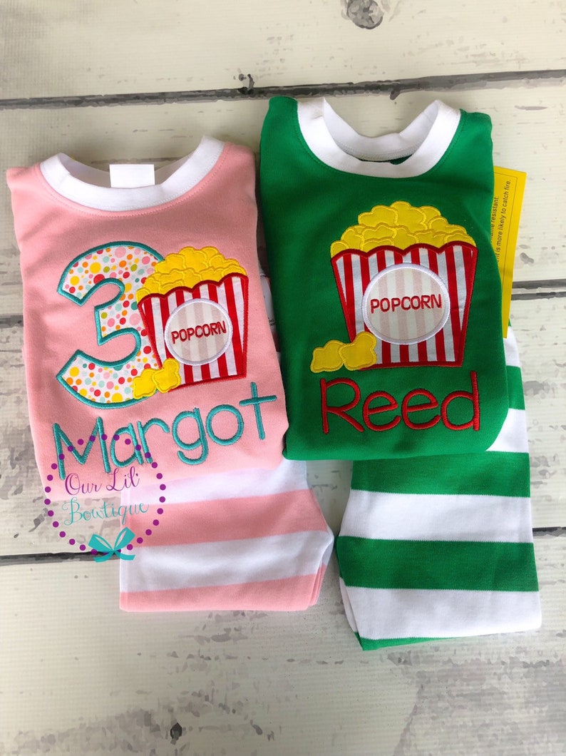 May include: Two pairs of pajamas with a popcorn theme. One pair is pink with white stripes and says "Margot" and "3". The other pair is green with white stripes and says "Reed".