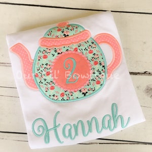 May include: A white t-shirt with a teal and pink embroidered teapot design. The teapot has a floral pattern and a circle with the number "2" in the center. The name "Hannah" is embroidered in teal below the teapot.