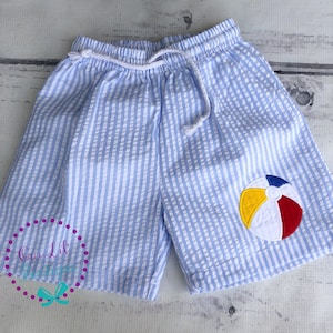 May include: Light blue and white seersucker shorts featuring an elastic waistband with a white drawstring cord. An embroidered beach ball applique with yellow, red, blue, and white segments is positioned on the lower right leg of the shorts.