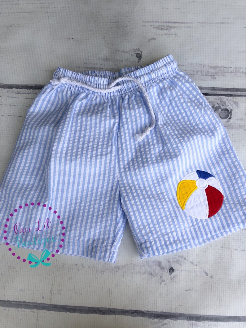 Puede incluir: Pantalones cortos de seersucker en azul claro y blanco con cintura el&aacute;stica y cord&oacute;n blanco. Una aplicaci&oacute;n bordada de una pelota de playa en amarillo, rojo, azul y blanco se encuentra en la pierna izquierda de la prenda.