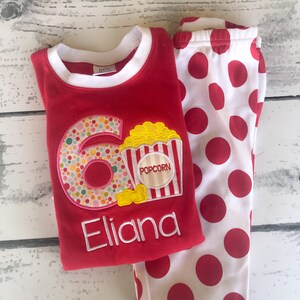 May include: Red and white polka dot pajamas with a red long-sleeved shirt featuring a colorful number six with popcorn and the name "Eliana" embroidered on it.