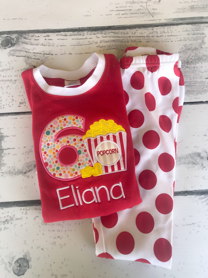 May include: Red and white polka dot pajamas with a red long-sleeved shirt featuring a colorful number six with popcorn and the name "Eliana" embroidered on it.