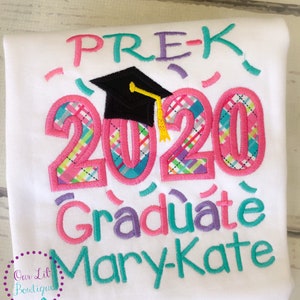 May include: White t-shirt with pink, blue, and purple embroidered text that reads "Pre-K 2020 Graduate Mary-Kate". The text is decorated with a black graduation cap and confetti.