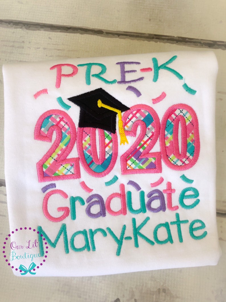 May include: White t-shirt with pink, blue, and purple embroidered text that reads "Pre-K 2020 Graduate Mary-Kate". The text is decorated with a black graduation cap and confetti.