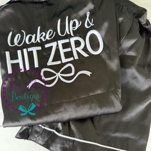 May include: Dark gray satin pajama set with white trim. The top features the embroidered text "Wake Up & HIT ZERO" in white, along with a bow design. The bottom is a matching pair of pajama pants. The set is from "Cute Boutique".