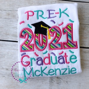 May include: White t-shirt with colorful embroidered text that reads "PRE-K 2021 Graduate McKenzie". The text is decorated with a graduation cap and confetti.