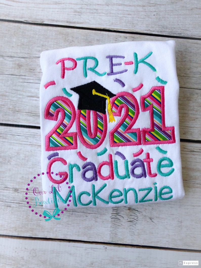 May include: White t-shirt with colorful embroidered text that reads "PRE-K 2021 Graduate McKenzie". The text is decorated with a graduation cap and confetti.