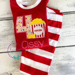 May include: Red and white striped pajamas with a red long-sleeved shirt featuring a large number four with colorful polka dots and a popcorn bucket with the word "POPCORN" on it. The shirt also has the name "Cissy" embroidered on it.