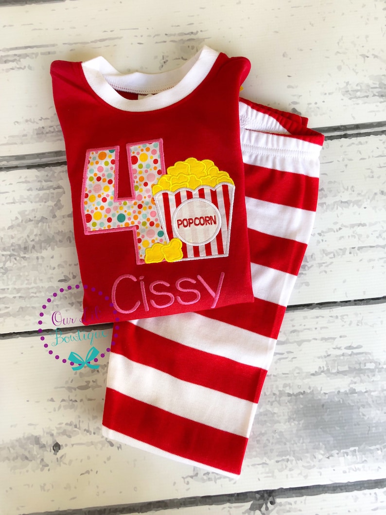 May include: Red and white striped pajamas with a red long-sleeved shirt featuring a large number four with colorful polka dots and a popcorn bucket with the word "POPCORN" on it. The shirt also has the name "Cissy" embroidered on it.