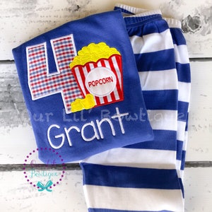 May include: Blue and white striped pants and a blue shirt with a red, white, and yellow popcorn applique and a blue and white gingham number four. The shirt also has the name "Grant" embroidered in white.