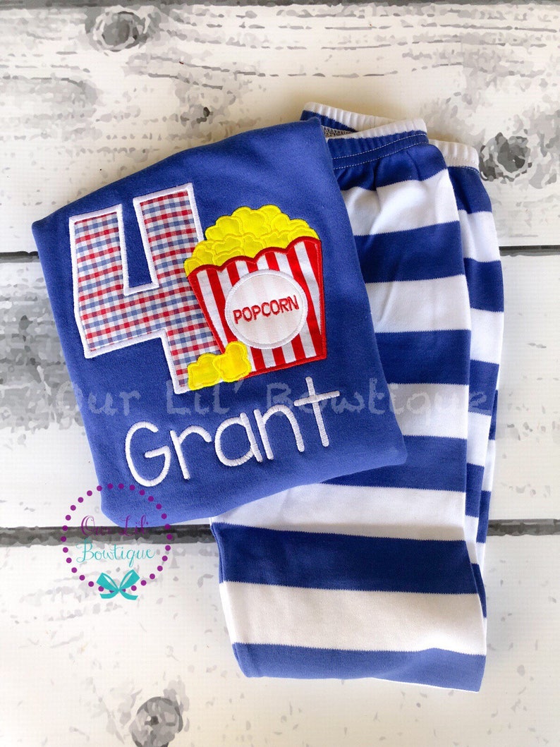 May include: Blue and white striped pants and a blue shirt with a red, white, and yellow popcorn applique and a blue and white gingham number four. The shirt also has the name "Grant" embroidered in white.