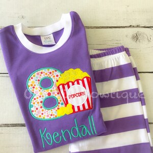 May include: Purple and white striped pajamas with a purple shirt featuring a large number 8 with colorful polka dots and a popcorn bucket with the word "POPCORN" on it. The shirt also has the name "Kendall" embroidered on it.