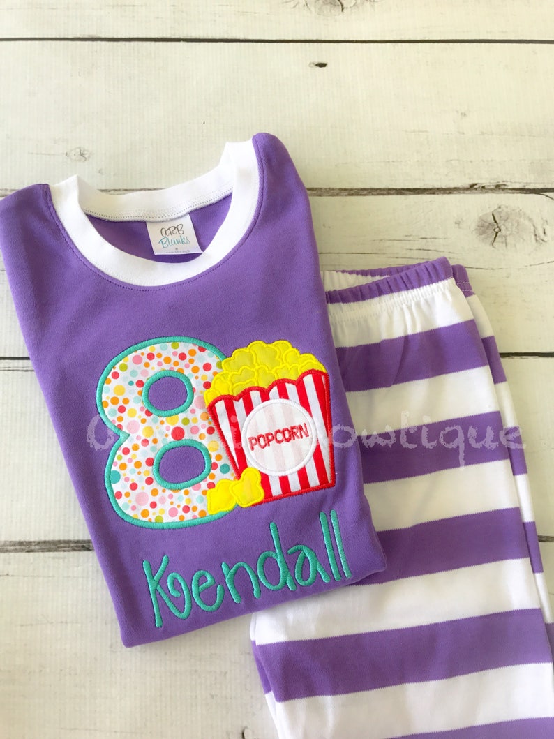 May include: Purple and white striped pajamas with a purple shirt featuring a large number 8 with colorful polka dots and a popcorn bucket with the word "POPCORN" on it. The shirt also has the name "Kendall" embroidered on it.