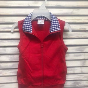 May include: A sleeveless red vest with a full-length zipper and a blue and white checkered collar. The vest is hanging on a white plastic hanger against a white slatted background. The vest appears to be made of a soft, knit fabric.