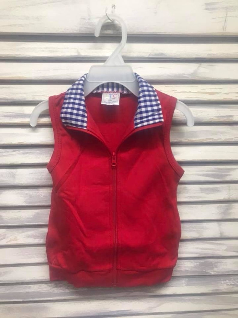 May include: A sleeveless red vest with a full-length zipper and a blue and white checkered collar. The vest is hanging on a white plastic hanger against a white slatted background. The vest appears to be made of a soft, knit fabric.