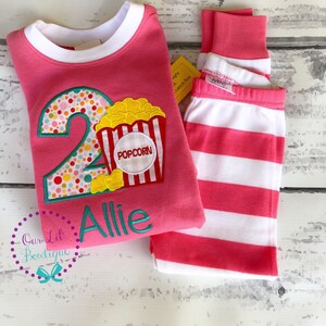 May include: Pink and white striped pajamas with a pink long-sleeved shirt featuring a large number 2 with colorful polka dots and a popcorn bucket with the word "Popcorn" on it. The shirt also has the name "Allie" embroidered on it.