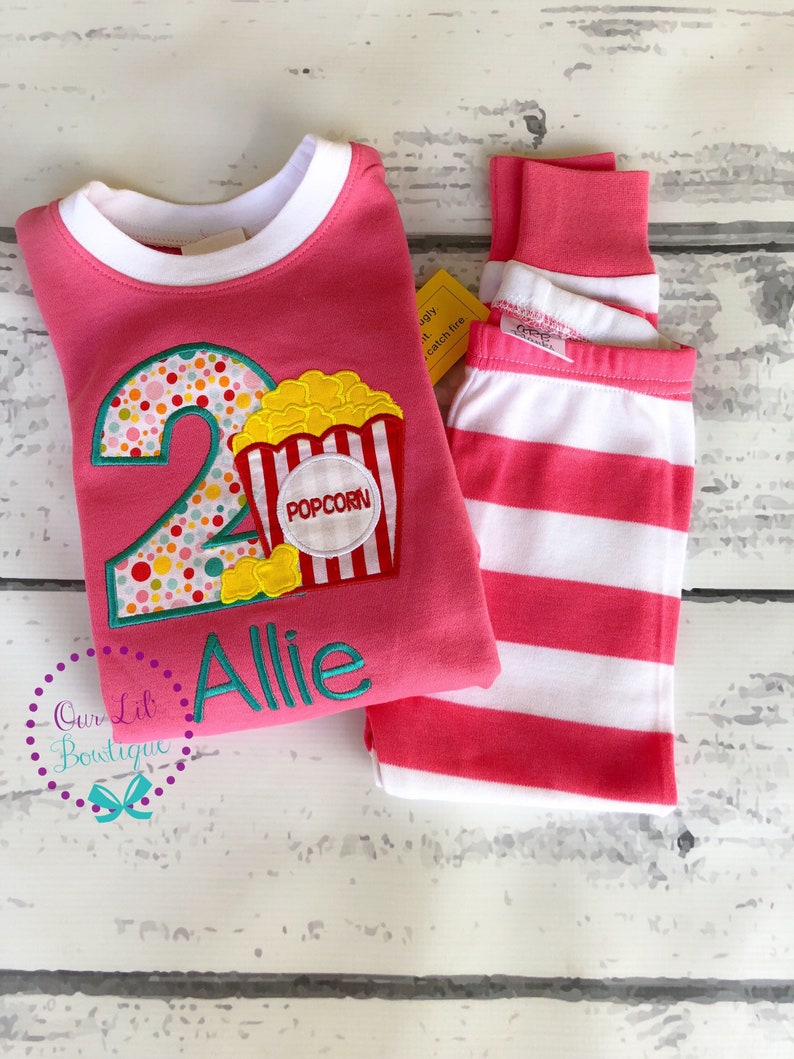May include: Pink and white striped pajamas with a pink long-sleeved shirt featuring a large number 2 with colorful polka dots and a popcorn bucket with the word "Popcorn" on it. The shirt also has the name "Allie" embroidered on it.
