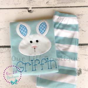 K&ouml;nnte beinhalten: Hellblaues Schlafanzug-Set mit einem Hasengesicht-Applikation, blau-wei&szlig; karierten Ohren und dem Namen "Griffin" in Blau gestickt. Das Set enth&auml;lt gestreifte Schlafanzughosen mit wei&szlig;en und hellblauen Streifen.