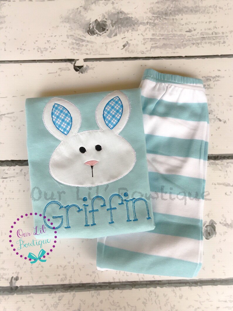 K&ouml;nnte beinhalten: Hellblaues Schlafanzug-Set mit einem Hasengesicht-Applikation, blau-wei&szlig; karierten Ohren und dem Namen "Griffin" in Blau gestickt. Das Set enth&auml;lt gestreifte Schlafanzughosen mit wei&szlig;en und hellblauen Streifen.