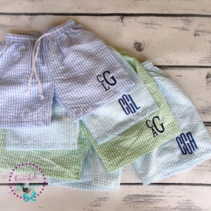Puede incluir: Una colecci&oacute;n de pantalones cortos de seersucker con patrones de cuadros vichy en azul claro y verde claro. Cada par tiene una cintura el&aacute;stica con cord&oacute;n blanco y un monograma bordado en azul oscuro en la pierna. El texto Our Lil Boutique es visible.