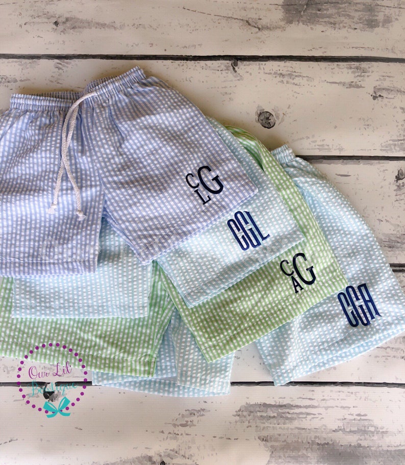Puede incluir: Una colecci&oacute;n de pantalones cortos de seersucker con patrones de cuadros vichy en azul claro y verde claro. Cada par tiene una cintura el&aacute;stica con cord&oacute;n blanco y un monograma bordado en azul oscuro en la pierna. El texto Our Lil Boutique es visible.