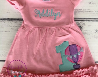 May include: A light pink short-sleeved dress with a ruffled hem. The chest features the embroidered name Addilyn in teal. The skirt displays a large teal number 1 appliqué next to a hot air balloon graphic with purple and pink accents.