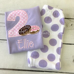 May include: A purple shirt with a pink polka dot number 2 and two donuts. The shirt says "Ellie" below the number. The shirt is paired with white leggings with purple polka dots.