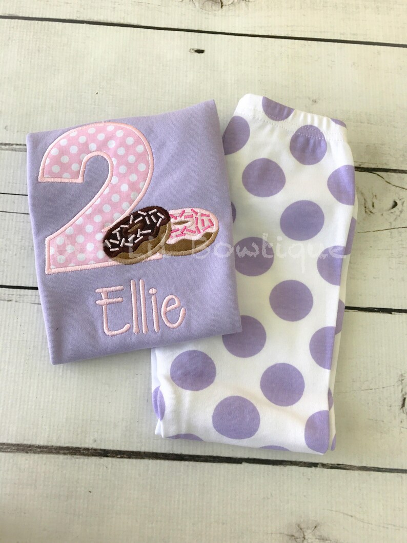 May include: A purple shirt with a pink polka dot number 2 and two donuts. The shirt says "Ellie" below the number. The shirt is paired with white leggings with purple polka dots.