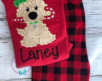 May include: A red long-sleeve shirt featuring an embroidered tan curly-haired dog with a green bow and a candy cane, with the name Laney embroidered in black below. Paired with red and black buffalo plaid pants with an elastic waistband.