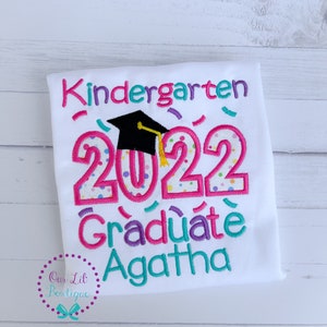 May include: White t-shirt with pink, blue, and green embroidery that says "Kindergarten 2022 Graduate Agatha". The number 2022 has a graduation cap on top of it.