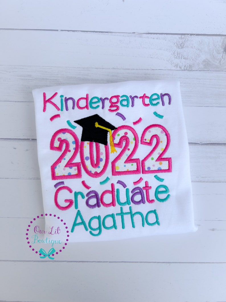 May include: White t-shirt with pink, blue, and green embroidery that says "Kindergarten 2022 Graduate Agatha". The number 2022 has a graduation cap on top of it.