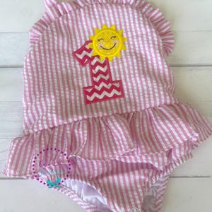 May include: Pink and white striped one-piece swimsuit with a ruffled neckline and hem. The swimsuit features a yellow sun applique and a pink number one with a zigzag design, perfect for a first birthday celebration.