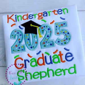 May include: White t-shirt with colorful embroidery that says "Kindergarten Graduate 2025" with a graduation cap and confetti. The name "Shepherd" is embroidered in green below.