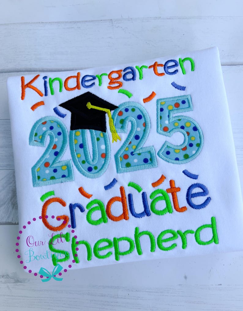 May include: White t-shirt with colorful embroidery that says "Kindergarten Graduate 2025" with a graduation cap and confetti. The name "Shepherd" is embroidered in green below.