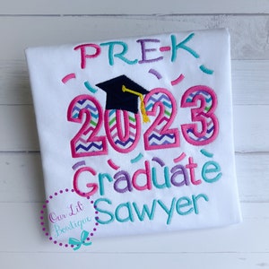 May include: White t-shirt with pink, purple, and teal embroidery. The text reads "PRE-K 2023 Graduate Sawyer" with a black graduation cap above the year.