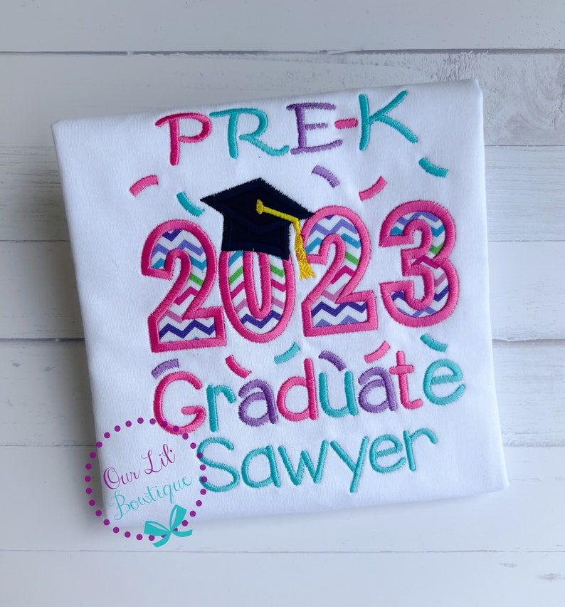 May include: White t-shirt with pink, purple, and teal embroidery. The text reads "PRE-K 2023 Graduate Sawyer" with a black graduation cap above the year.