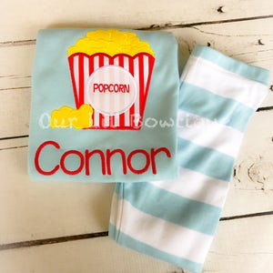 May include: A light blue long-sleeved shirt with a red and white striped popcorn bucket design. The shirt has the word "POPCORN" in a circle on the bucket and the name "Connor" embroidered below the bucket. The shirt is paired with light blue and white striped pants.