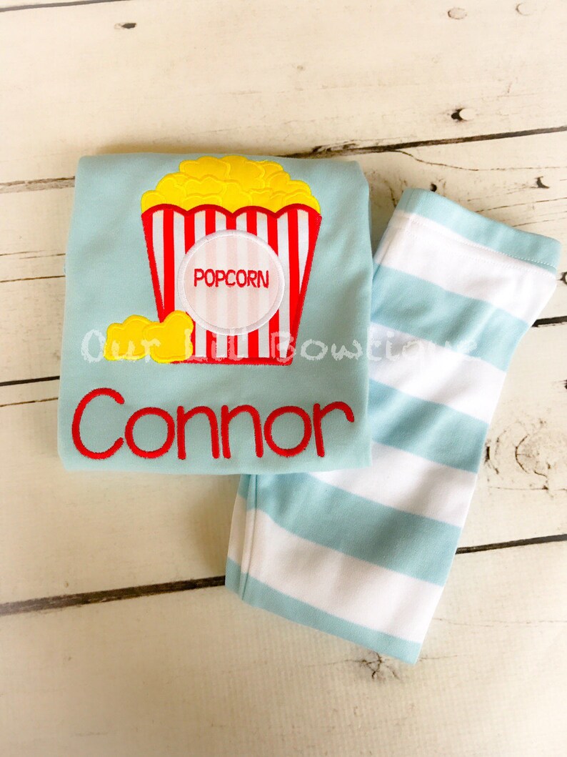 May include: A light blue long-sleeved shirt with a red and white striped popcorn bucket design. The shirt has the word "POPCORN" in a circle on the bucket and the name "Connor" embroidered below the bucket. The shirt is paired with light blue and white striped pants.