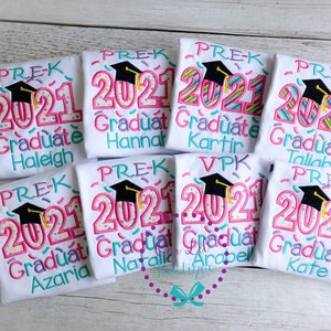 May include: Eight white t-shirts with colorful embroidered designs. Each shirt says "PRE-K 2021 Graduate" with a graduation cap and a different name underneath. The names are Haleigh, Hannah, Kartir, Taliah, Azaria, Natalie, Arobell, and Kate.
