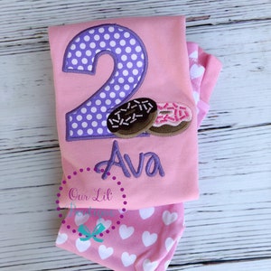 May include: Pink and purple two-piece outfit with a donut design and the name Ava embroidered on the shirt.