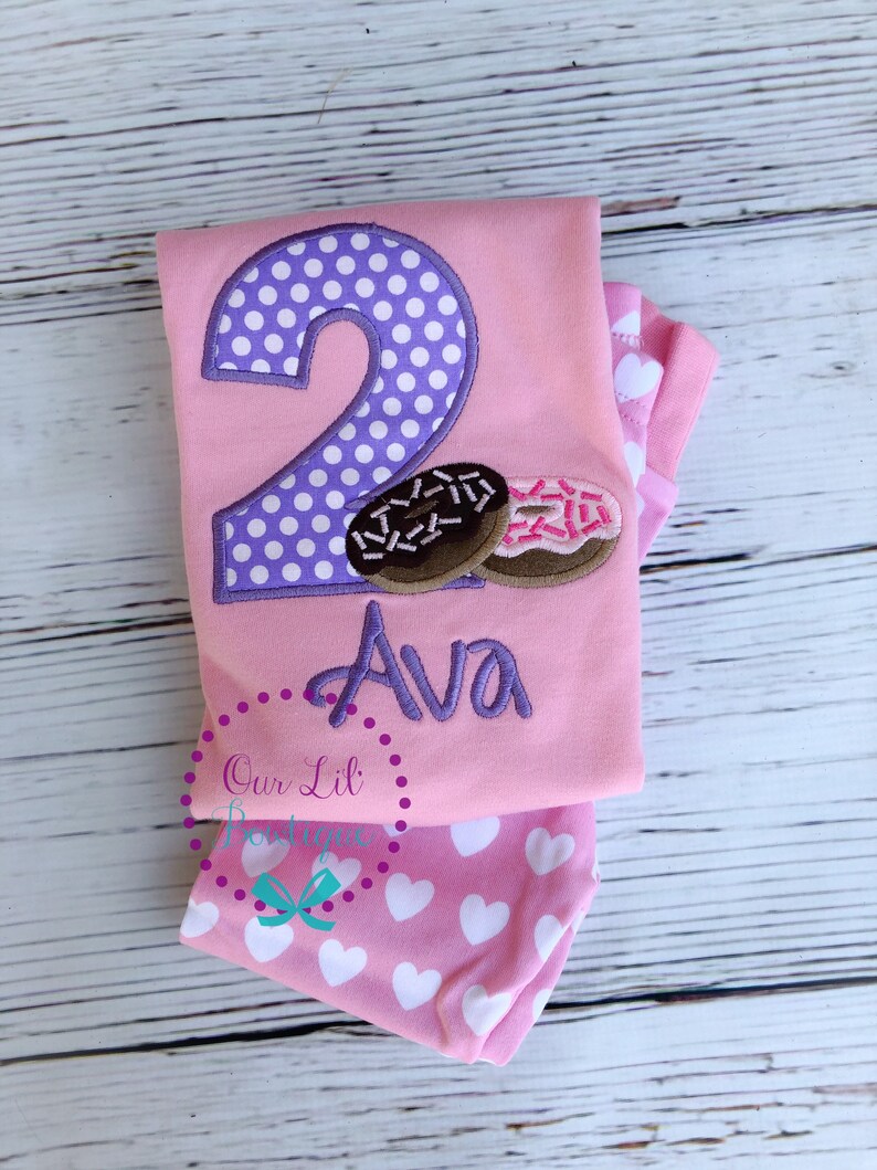 May include: Pink and purple two-piece outfit with a donut design and the name Ava embroidered on the shirt.