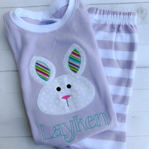 K&ouml;nnte beinhalten: Ein lavendelfarbenes Schlafanzug-Set mit einem Hasen-Applikation mit bunten gestreiften Ohren und einer rosa Nase. Das Shirt hat den Namen "Layken" in T&uuml;rkis gestickt. Die passende Hose ist in Lavendel und Wei&szlig; gestreift.