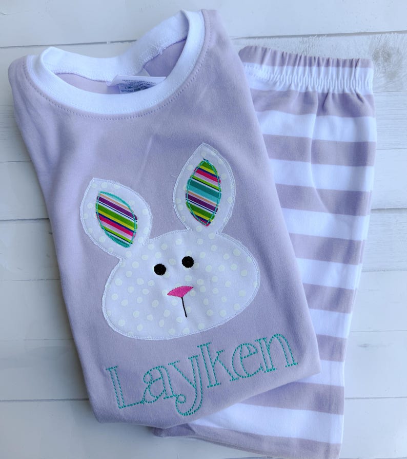 K&ouml;nnte beinhalten: Ein lavendelfarbenes Schlafanzug-Set mit einem Hasen-Applikation mit bunten gestreiften Ohren und einer rosa Nase. Das Shirt hat den Namen "Layken" in T&uuml;rkis gestickt. Die passende Hose ist in Lavendel und Wei&szlig; gestreift.