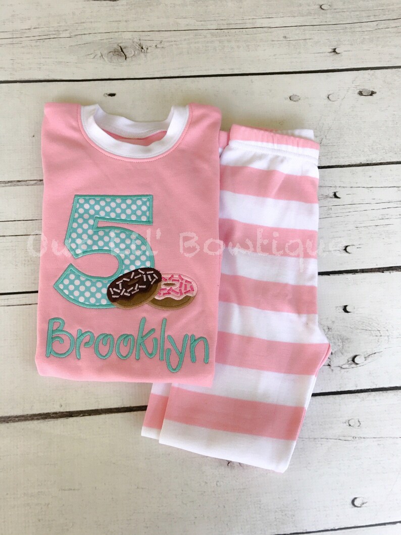 May include: Pink and white striped pajamas with a pink shirt featuring a turquoise number 5 with white polka dots and two donuts. The shirt also has the name "Brooklyn" embroidered in turquoise.