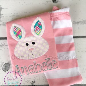 K&ouml;nnte beinhalten: Ein rosa Baby-Outfit mit einem Hasengesicht-Design, Tupfen und Karodetails. Das Outfit beinhaltet ein Oberteil mit dem Namen "Anabelle" in T&uuml;rkis gestickt und eine Hose mit rosa und wei&szlig;en Streifen. Das Logo "Our Lil Boutique" ist ebenfalls sichtbar.