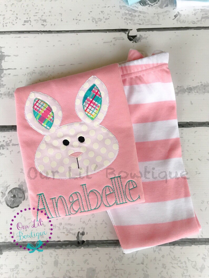 K&ouml;nnte beinhalten: Ein rosa Baby-Outfit mit einem Hasengesicht-Design, Tupfen und Karodetails. Das Outfit beinhaltet ein Oberteil mit dem Namen "Anabelle" in T&uuml;rkis gestickt und eine Hose mit rosa und wei&szlig;en Streifen. Das Logo "Our Lil Boutique" ist ebenfalls sichtbar.