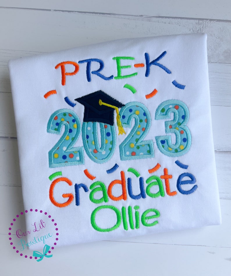 May include: White t-shirt with blue, green, and orange embroidery that says "PRE-K 2023 Graduate Ollie". A blue graduation cap is embroidered above the year.